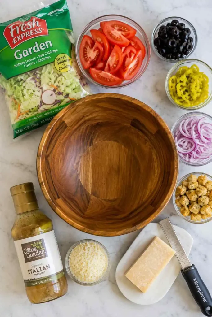 Authentic Copycat Olive Garden Salad Recipe (15-Minute Meal) 2 Flatlay view of bagged salad mix, sliced tomatoes, pepperoncini, black olives, red onions, croutons, and Parmesan cheese arranged in glass bowls.