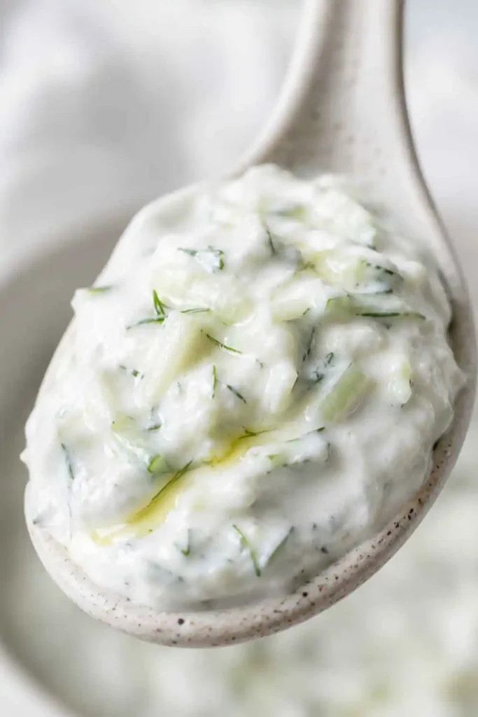 Close-up of thick creamy tzatziki sauce texture on a ceramic spoon showing cucumber flecks and fresh dill