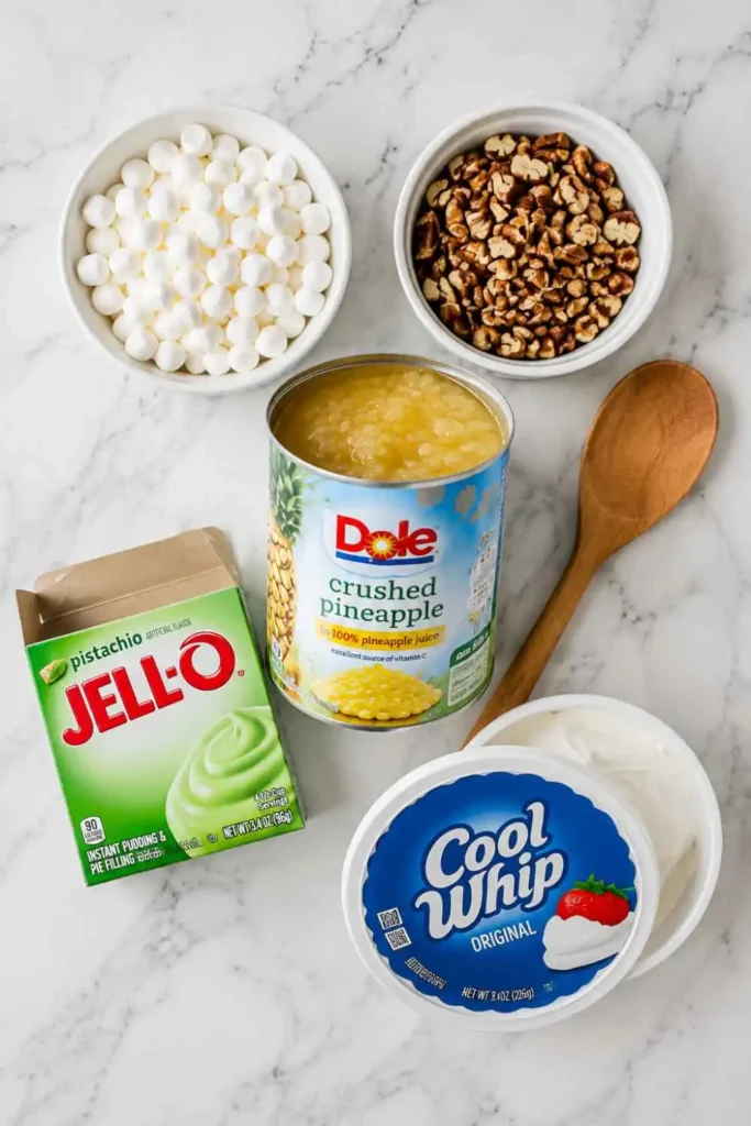 All 5 ingredients for Watergate salad recipe arranged on white marble counter including pistachio pudding, pineapple, marshmallows, Cool Whip, and pecans
